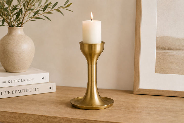 A brass candle holder with a lit white pillar candle, styled on a wooden table next to a vase with greenery and a framed print.