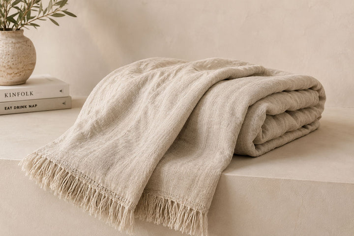 A folded beige linen throw blanket with fringed edges draped over a light stone surface, with a small vase of greenery and stacked books in the background.