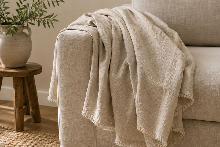 A light beige textured throw blanket with fringed edges draped over the arm of a matching beige sofa in a cozy living room setting, with a small wooden side table and potted plant visible on the left.